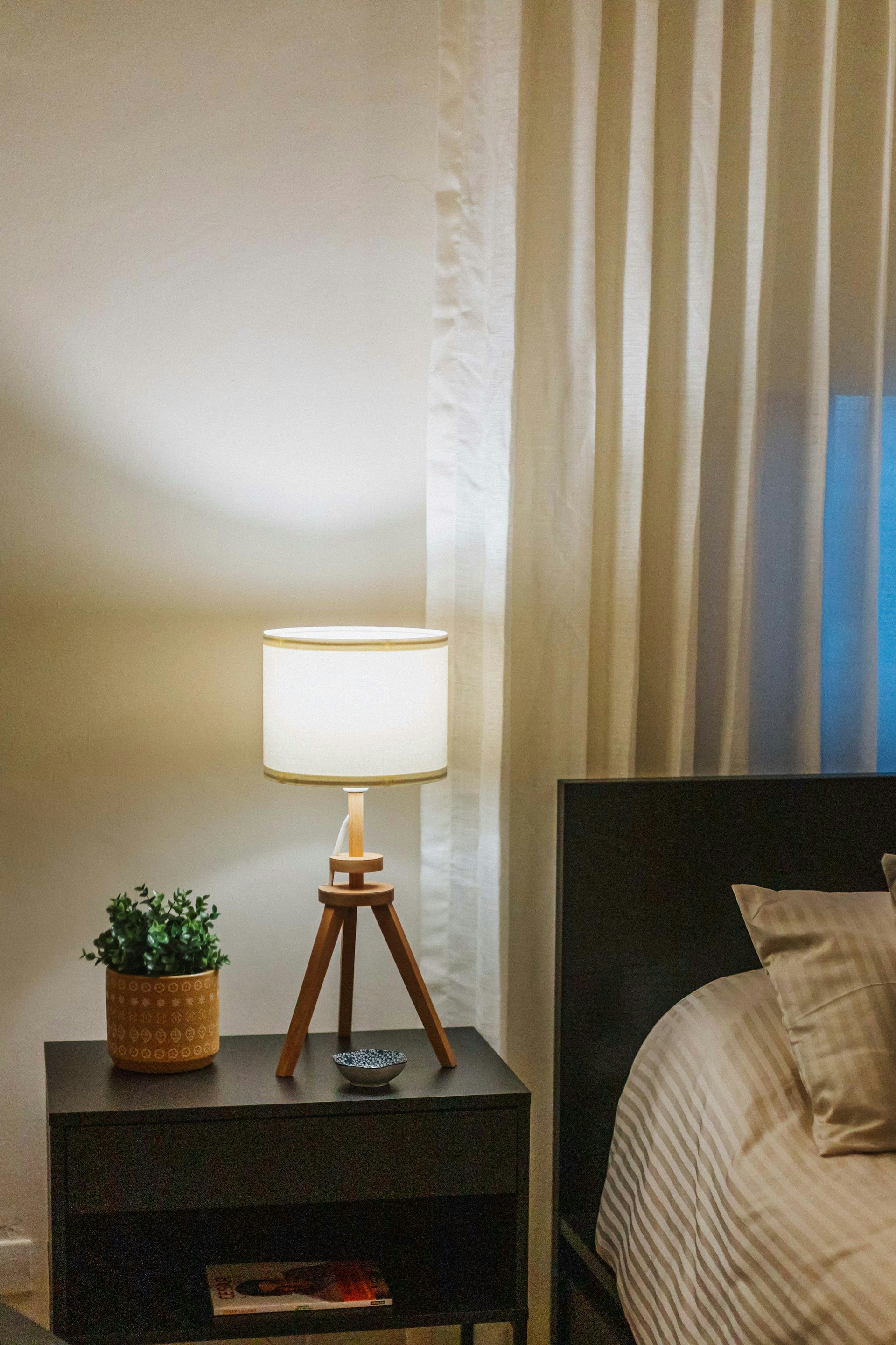 A tripod lamp with a white shade glowing beside linen curtains