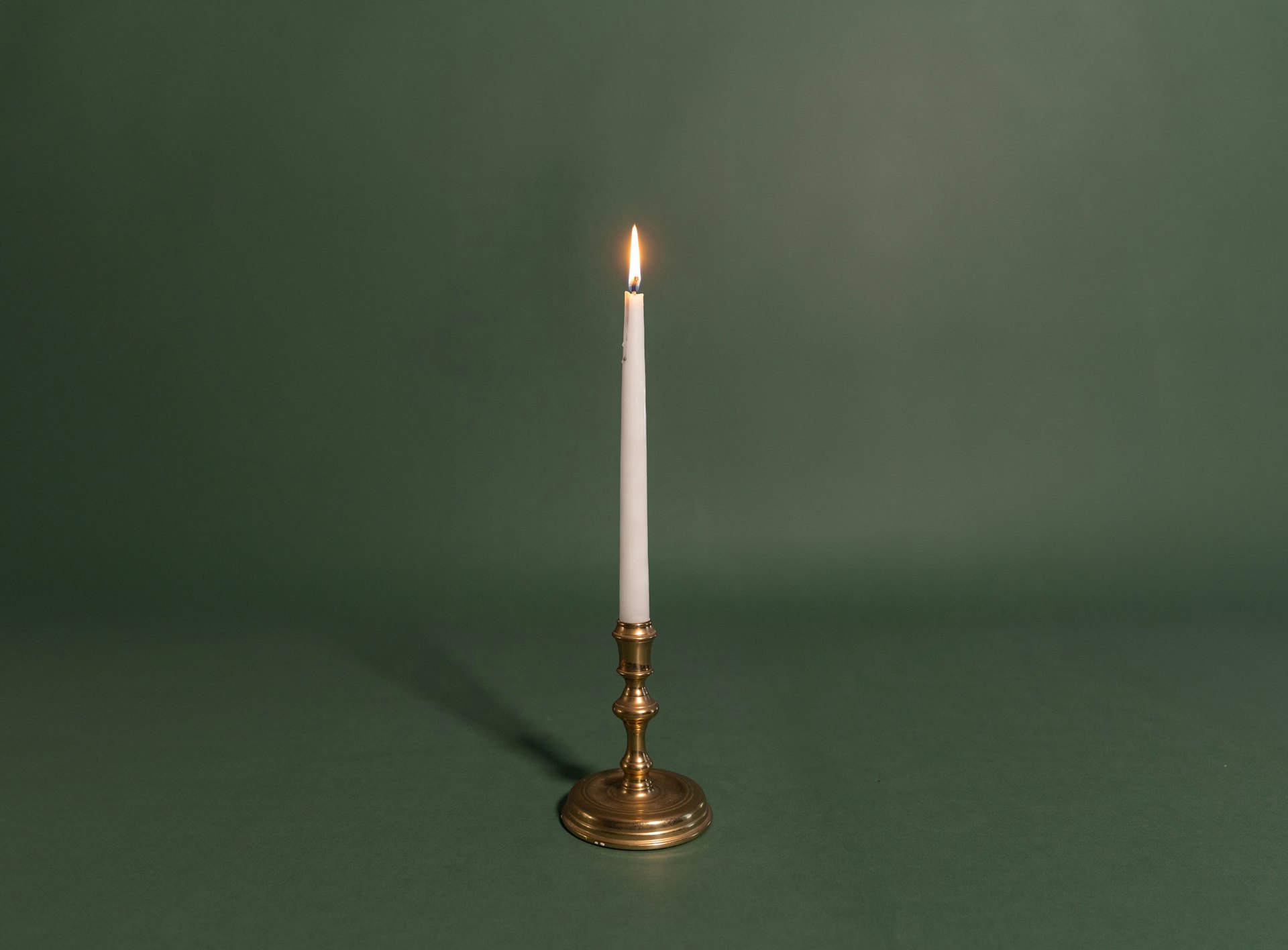 A single white taper candle in a slim brass holder against a green wall